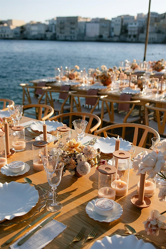 Reception tablescape with white scalloped plates, menus, glassware and taper candles on a wood table, with waterfront skyline behind