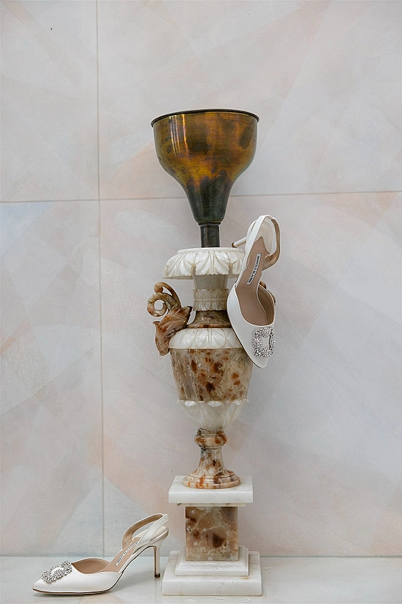 Bridal shoes in a wedding shoe flatlay, white pointed-toe slingback heels with rhinestone buckle on a marble pedestal by a decorative urn