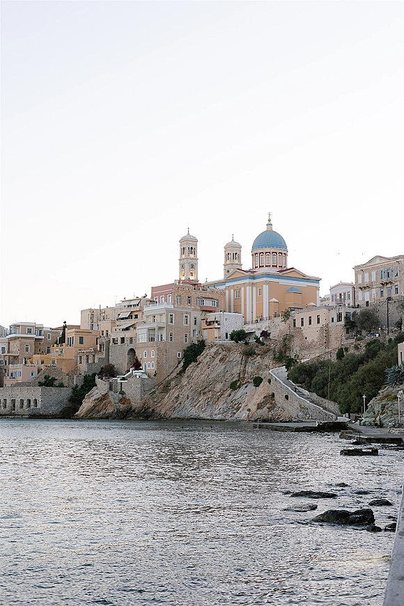 Coastal church view with a blue dome above stone buildings, overlooking sea and rocky shoreline along hillside cliffs and promenade