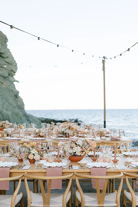 Reception tablescape with an outdoor reception table of wood farmhouse seating, pastel florals, taper candles, and string lights by the ocean cliffs