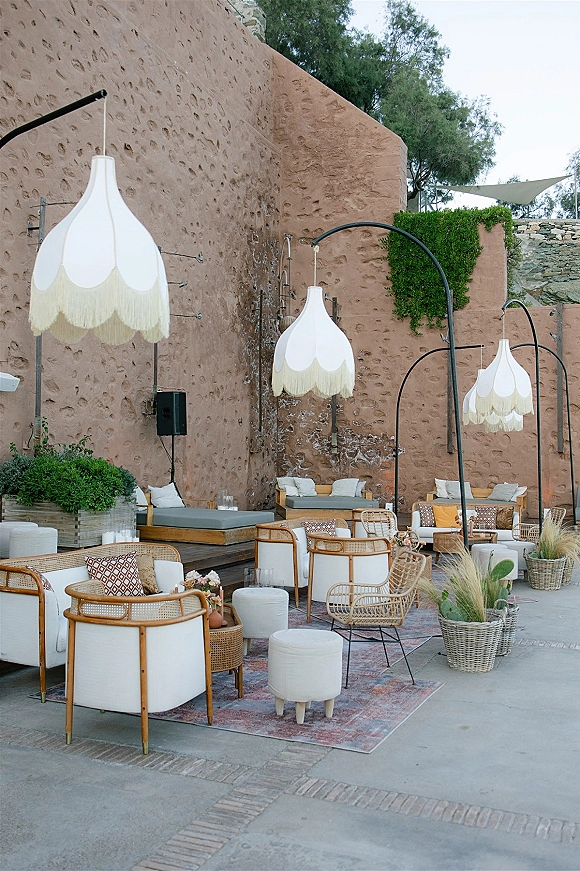 Wedding lounge area with rattan chairs, sofas and layered rugs, lit by hanging lanterns with candles and pampas grass in a courtyard