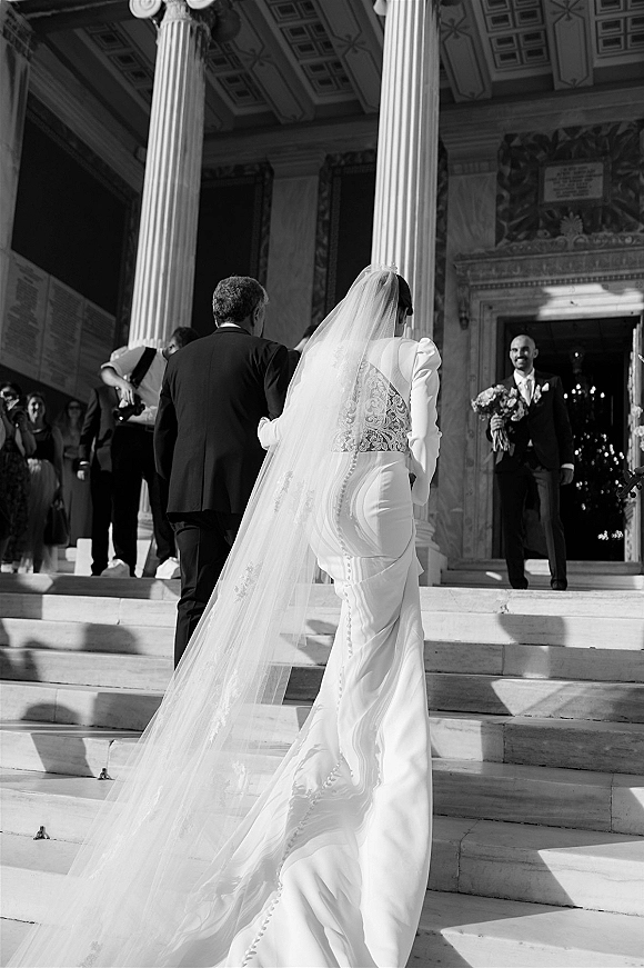 Wedding processional with bride walking up steps, long cathedral veil and train flowing as guests watch near grand columned entrance