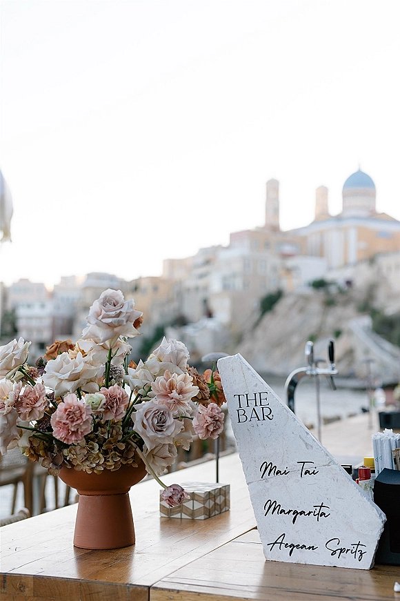 Wedding bar sign with a stone wedding bar menu and handwritten cocktail list on a wooden table with terracotta vase and florals, seaside terrace backdrop