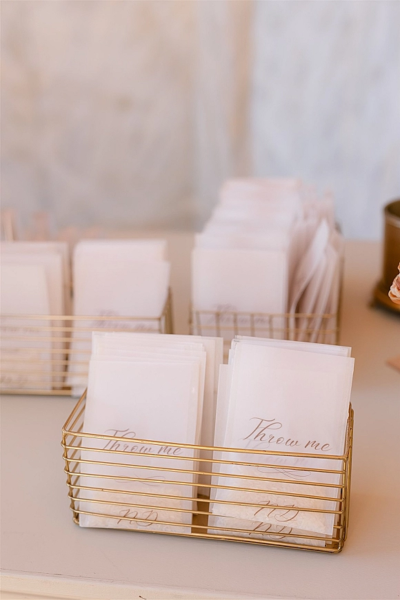 Wedding confetti packets arranged as a confetti toss idea in a gold wire basket on a white tabletop beside a candle and neutral wall