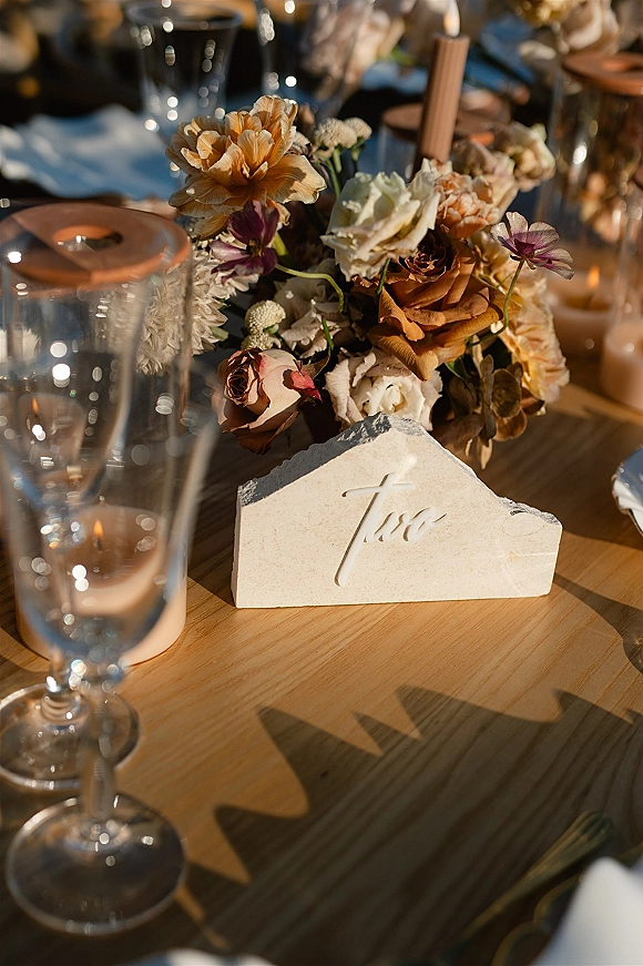 Reception tablescape with wedding table centerpiece of low rose florals, brown taper candle, votives, glassware, and stone place card on a wood table
