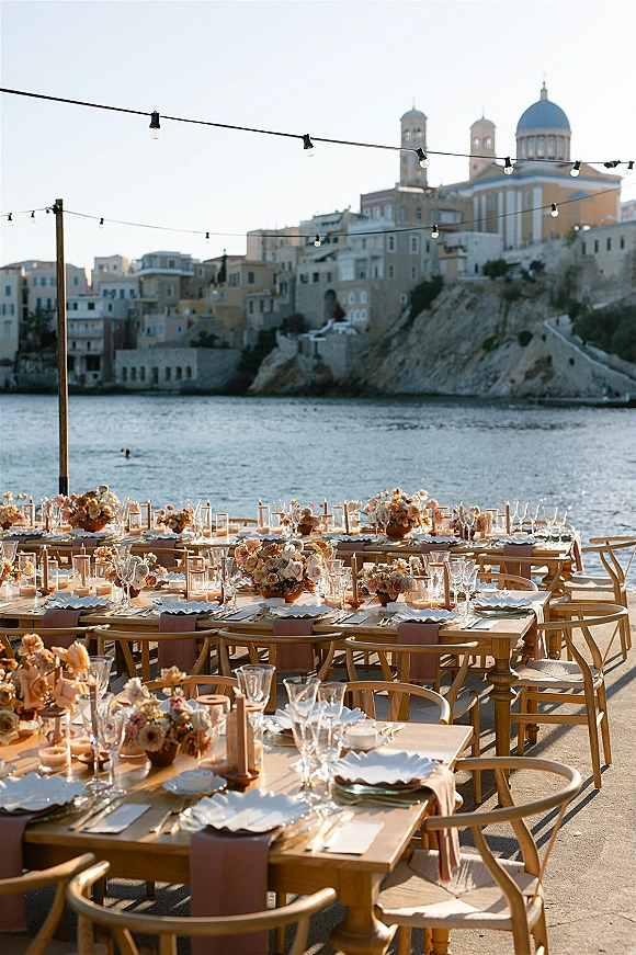 Outdoor reception tablescape with a long banquet table setup, floral centerpieces and taper candles under string lights by a waterfront town backdrop
