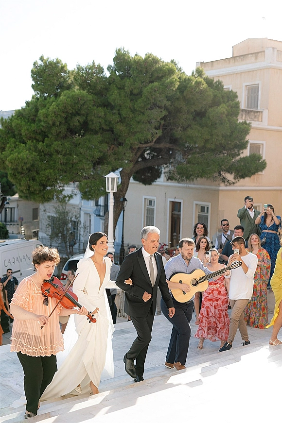 Wedding processional as bride in long sleeve dress and veil walks down outdoor steps past violinist and guitarist, guests watching in daylight