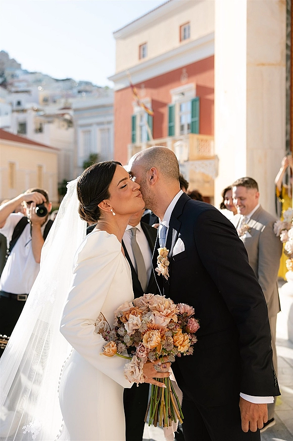 Wedding kiss as bride holds a peach and blush bouquet, veil flowing, while guests and a photographer line a sunlit street with buildings behind