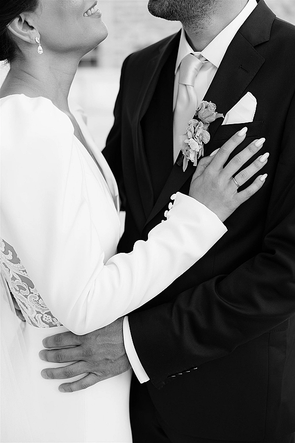 Couple portrait in a black and white wedding portrait, bride’s lace sleeve and ring on groom’s chest beside boutonniere, soft blurred wall backdrop