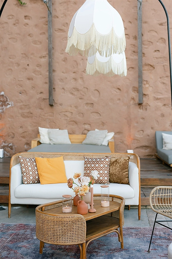 Wedding lounge area with rattan sofa and throw pillows, candles and florals on a coffee table under a fringe pendant lamp by a stucco wall