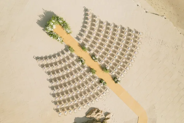 Beach ceremony setup with aerial wedding ceremony layout, white chairs lining a tan runner to a floral arch on sand by rocks