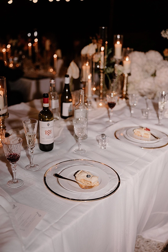 Reception tablescape with candlelit wedding table featuring white and gold place settings, hydrangeas, wine bottles, and string lights in dim light