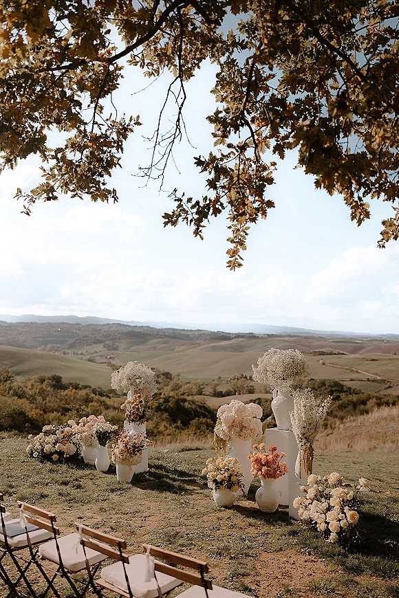Ceremony altar decor with floral arrangements on white pedestal plinths and vases, framed by aisle chairs and rolling hills backdrop