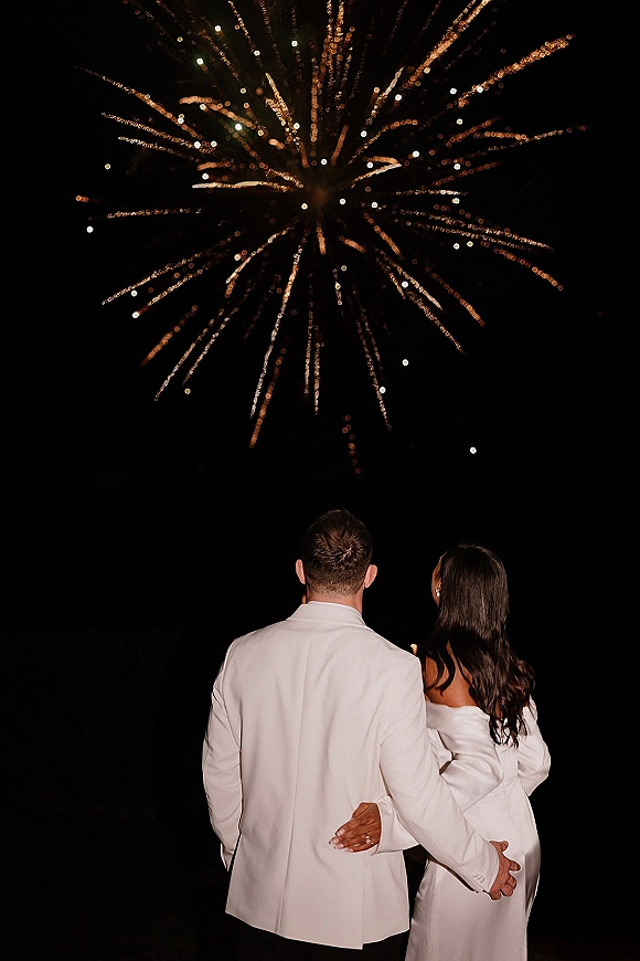 Fireworks wedding photo of newlyweds watching fireworks exit arm in arm, bride in white dress showing ring, under a night sky