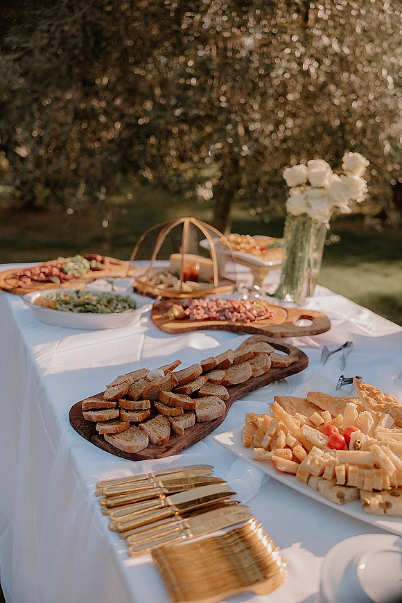 Cocktail hour food spread with a charcuterie board wedding display of bread, cheese and snacks on white tablecloth outdoors in sunlight