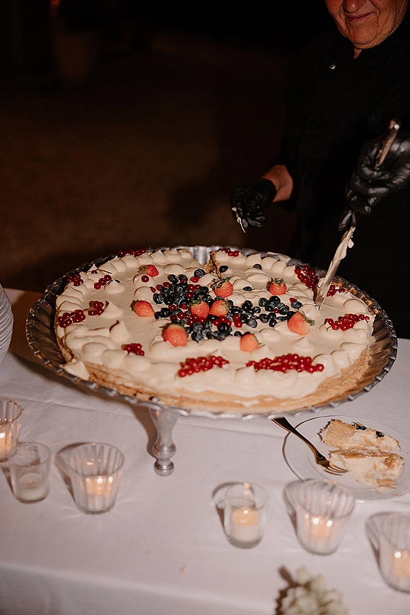 Wedding cake with berry wedding cake topping on a cake stand, with cake server, dessert plate, and votive candles on a reception table
