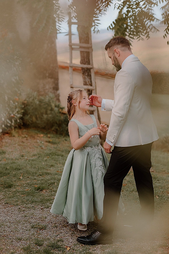Flower girl and groom sharing a sweet moment as he fixes her hair in a garden, wearing a white tuxedo jacket and boutonniere