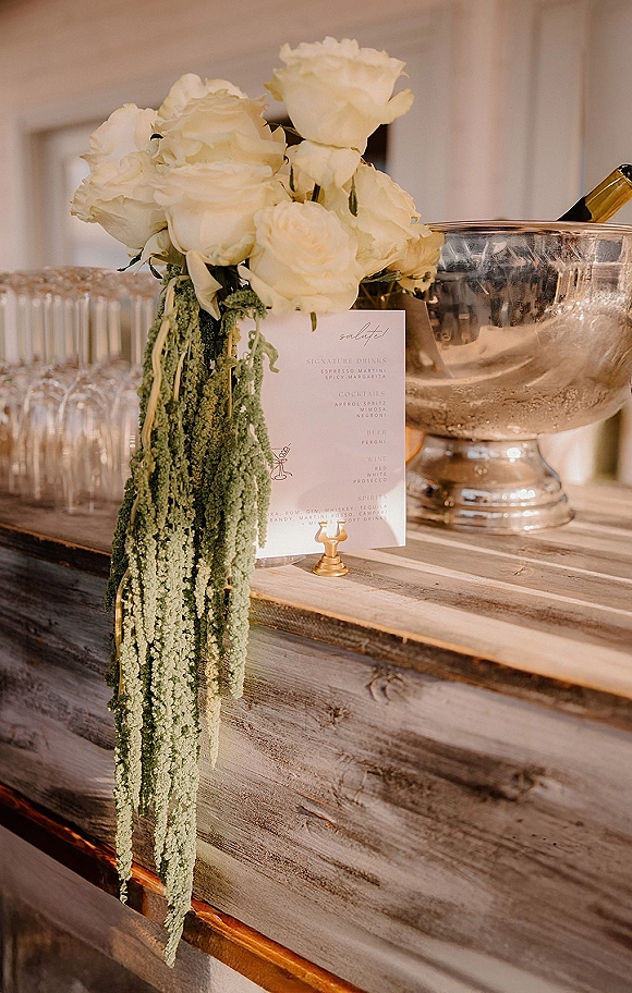 Wedding bar setup with a cocktail menu card in a brass holder, white rose arrangement, coupe glasses, and champagne on ice against a white paneled wall
