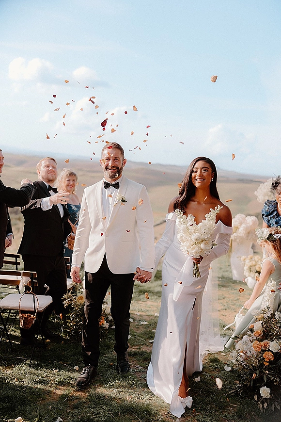 Wedding recessional as bride and groom walk the aisle holding hands through flower petals, bride with bouquet, hills and cloudy sky behind