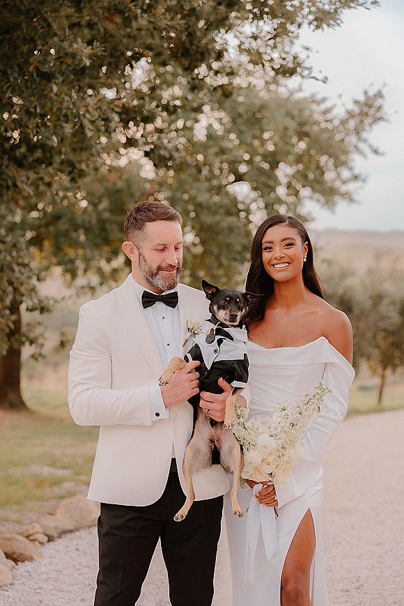 Couple portrait of bride and groom with dog, bride holding bouquet beside a dirt path with trees and open sky backdrop.