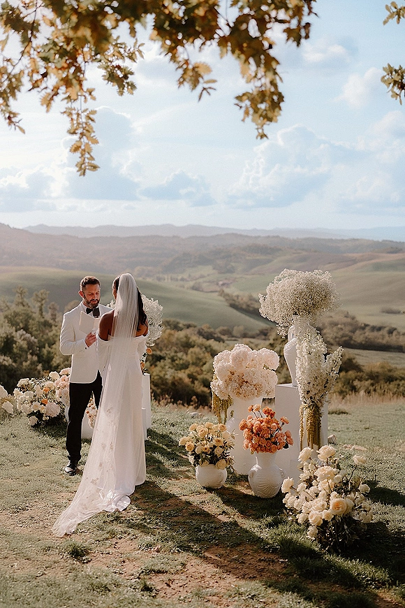 Wedding vows as the couple exchanging vows beside white pedestal florals, bride in veil and groom in tuxedo, with rolling hills beyond