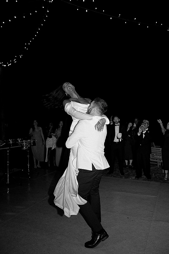 First dance moment on a wedding dance floor as the groom lifts the bride in her gown under string lights at a nighttime outdoor reception