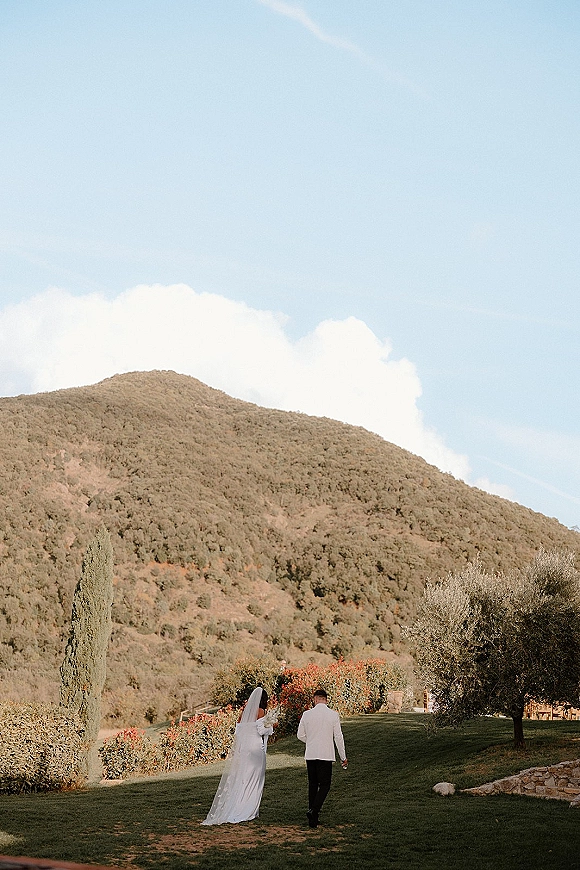 Couple walking away hand in hand, bride’s veil and dress train flowing beside groom’s suit on a mountain hillside under blue sky