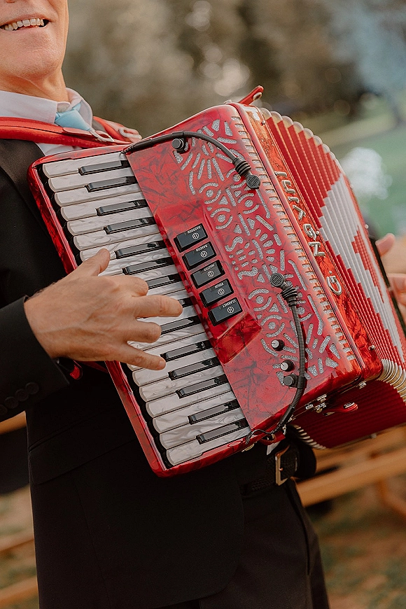 Wedding musician playing a red accordion with a lapel microphone, wearing a suit and tie near trees and a wooden fence outdoors