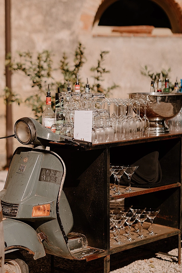 Wedding bar setup with a mobile wedding bar cart, glassware and liquor bottles beside a vintage scooter in a stone courtyard doorway