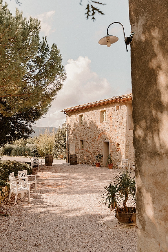 Outdoor wedding venue with white wooden chairs lining a gravel courtyard, wine barrels and potted plants beside a stone farmhouse backdrop
