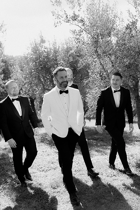 Groom with groomsmen walking photo across a sunlit lawn under trees, the groom in a white dinner jacket with black bow tie