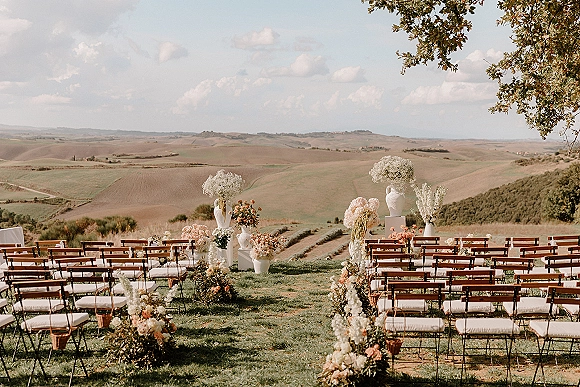 Ceremony setup for an outdoor wedding ceremony with wooden chairs and peach-white aisle florals in urns overlooking rolling farmland hills
