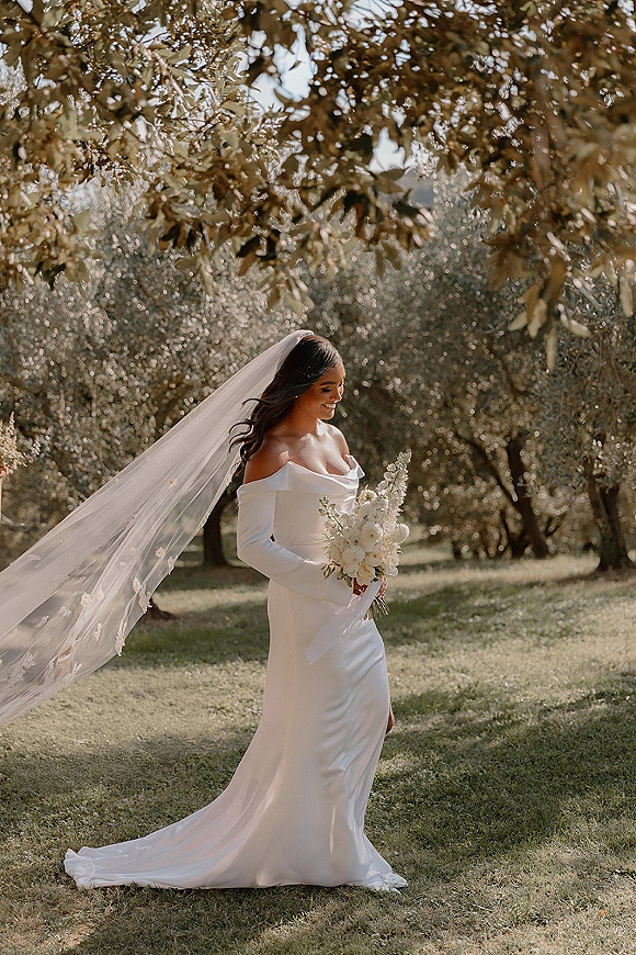Bridal portrait of a bride holding bouquet in an off the shoulder wedding dress, long veil flowing, on a sunlit lawn under trees