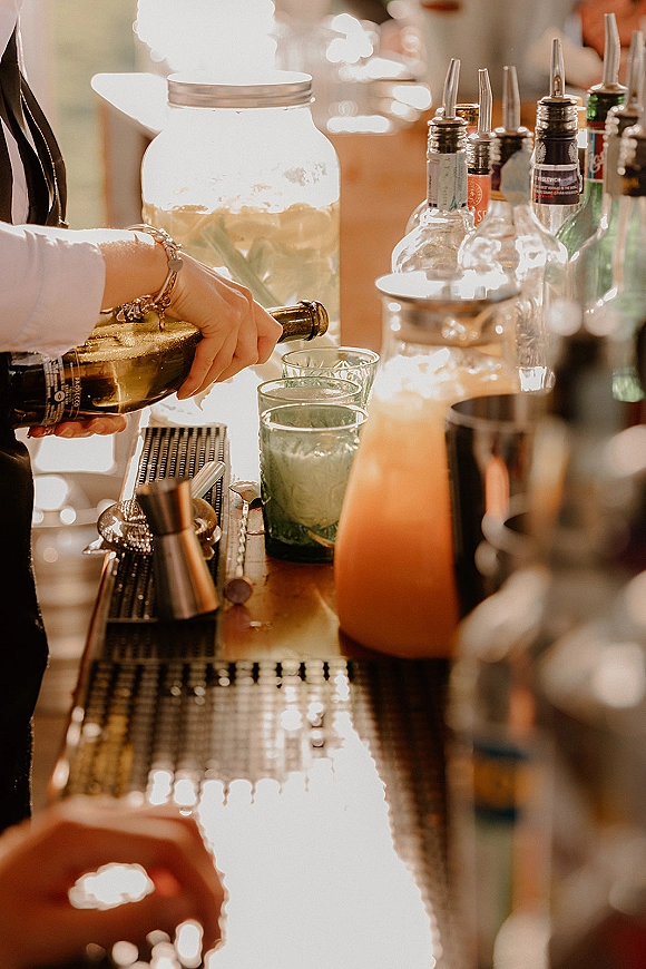 Wedding bar service setup with liquor bottles, cocktail shaker, and glass tumblers beside a large beverage dispenser on a warmly lit counter