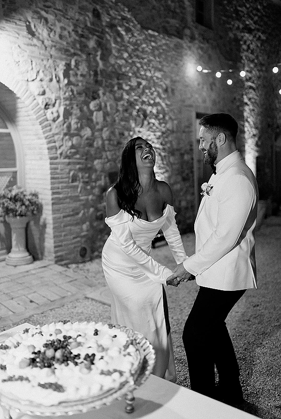 Couple portrait in a black and white wedding portrait, bride in off-shoulder gown and groom in tux laughing by cake under string lights in courtyard