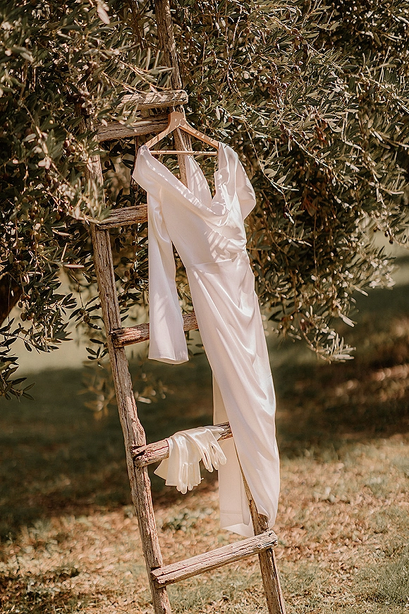 Wedding dress in silk wedding dress fabric hanging on a wooden hanger from a vintage ladder beside an olive tree on a lawn