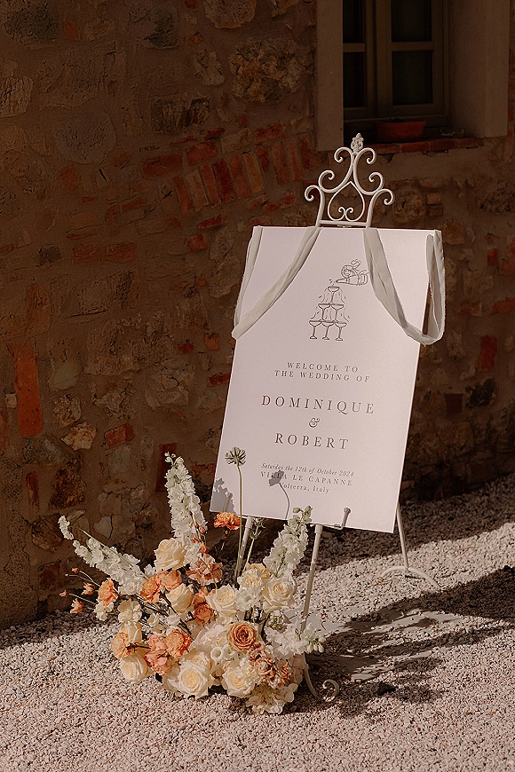 Wedding welcome sign on an ornate white metal easel with ribbon drape and rose, delphinium florals at the base by a stone wall window