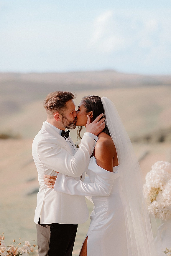 Wedding kiss portrait of bride and groom kissing, her long veil and off-shoulder dress beside his white tux, with desert mountains behind