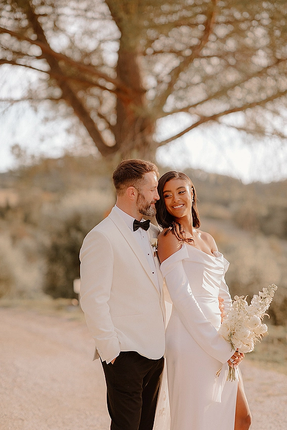 Couple portrait of bride and groom portrait at golden hour, bride in off-shoulder gown holding bouquet beside groom in white tuxedo under a large tree with hills behind
