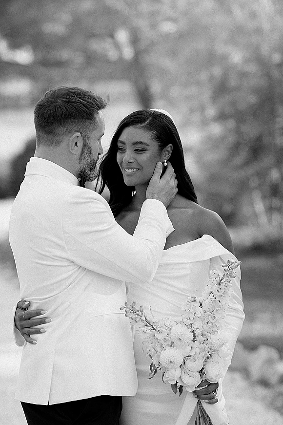 Couple portrait of bride and groom embrace as he touches her face, she holds a bouquet, in black and white with blurred trees behind