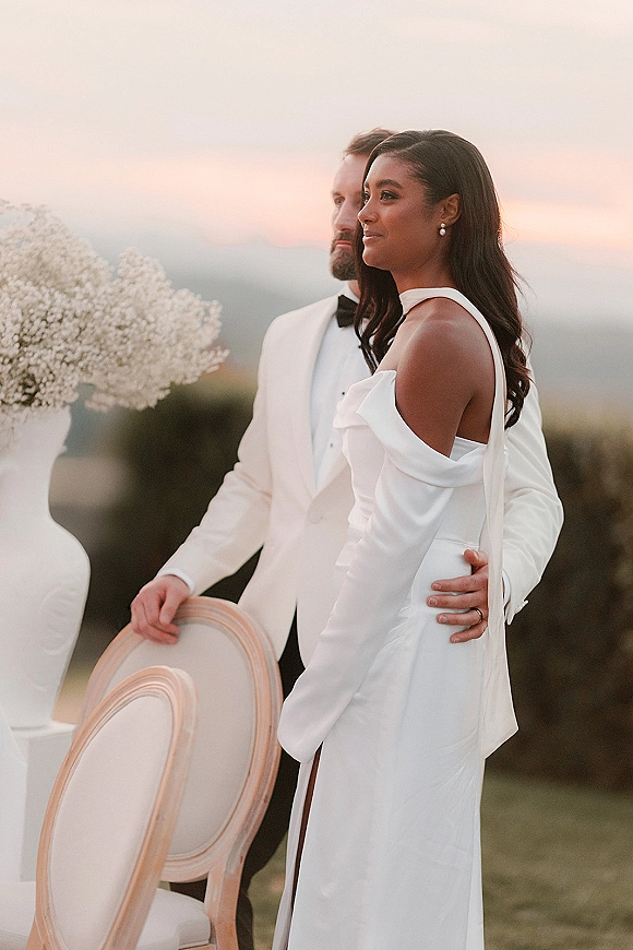 Couple portrait of bride and groom portrait in side profile, her off-shoulder gown and his white tuxedo at sunset hills backdrop