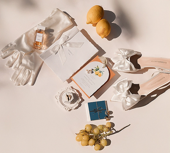 Wedding flat lay with wedding invitation suite, ribbon bow, rings, pearl earrings, perfume, satin gloves, shoes, and citrus on white surface