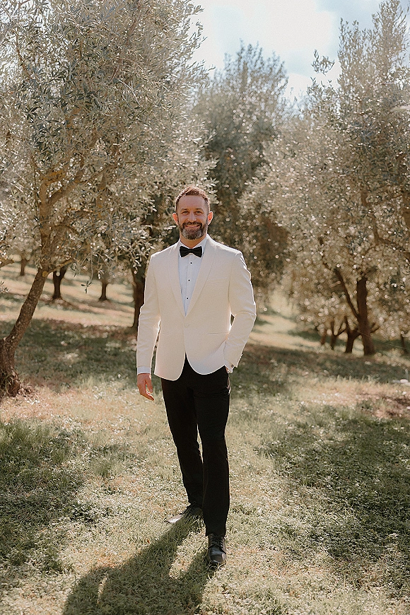 Groom portrait in a white tuxedo jacket and black bow tie, standing in sunlight among olive trees with a grassy field behind