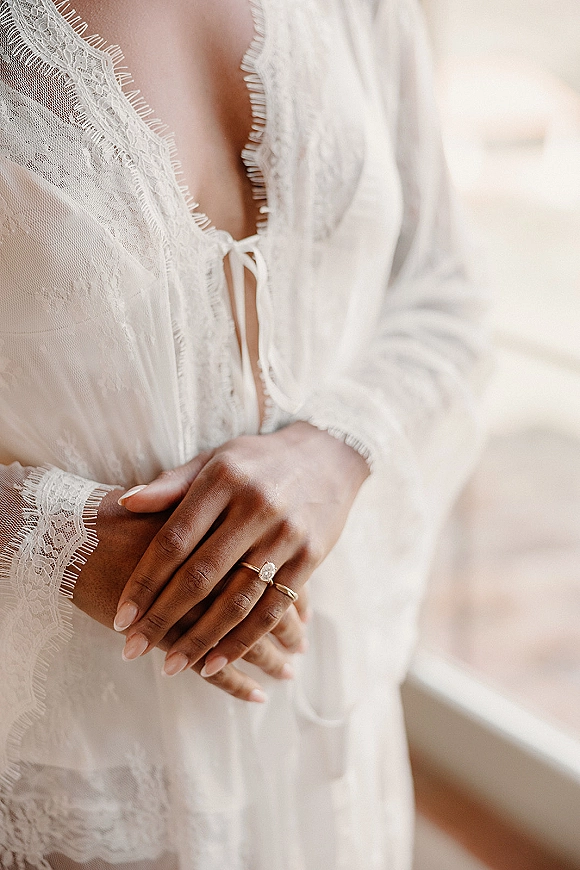 Engagement ring and oval diamond ring stacked with wedding band on nude-manicured hand, framed by lace robe and soft window light