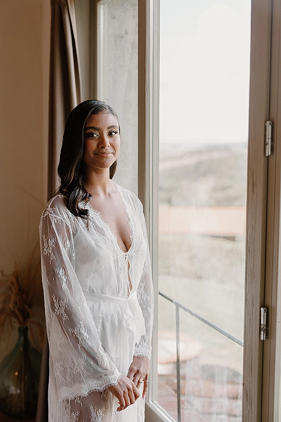 Bridal portrait of a bride by window in a lace bridal robe, showing her engagement ring in soft natural light with curtains behind