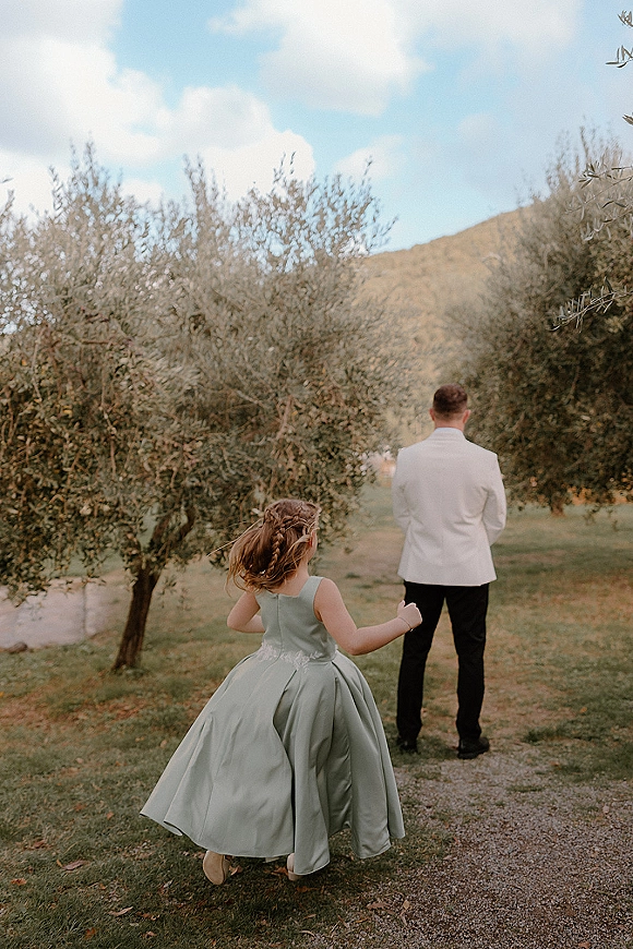 Wedding first look as a flower girl running toward a groom in a white dinner jacket on a tree-lined path with mountains under blue sky