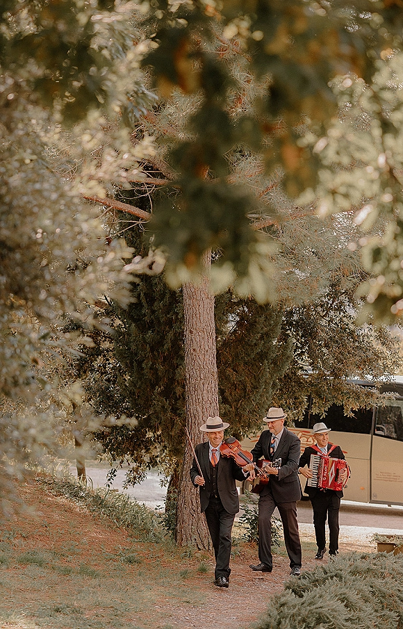 Wedding musicians in suits and ties walk down a dirt path playing violin, accordion, and guitar, with trees and a tour bus behind them