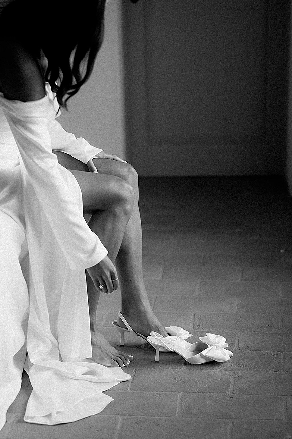 Bridal getting ready moment as bride putting on shoes, showing off-the-shoulder robe and engagement ring on a brick floor indoors