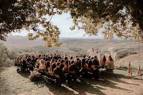 Outdoor wedding ceremony on a hillside with bride in long veil and groom in black suit at a floral altar under tree canopy