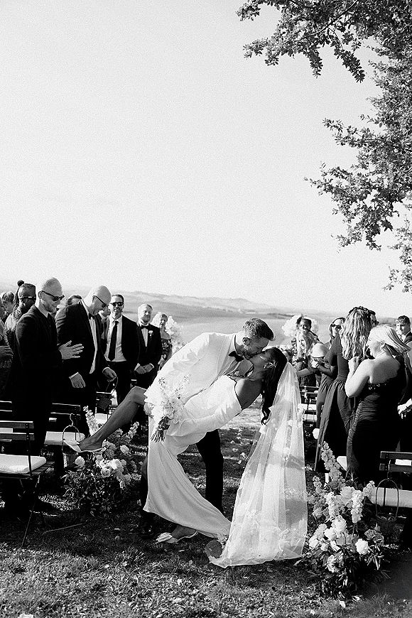 Wedding kiss moment as groom dips the bride, her veil and bouquet flowing along a flower-lined outdoor aisle with cheering guests and hills beyond